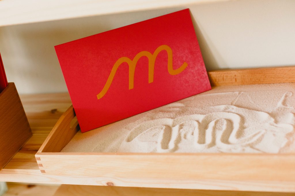Montessori cursive letter "M" and sand tray with letter written in sand. one way Montessori education works by teaching writing.