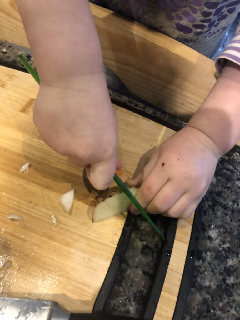 Toddler using plastic knife to cut potatoes for soup.