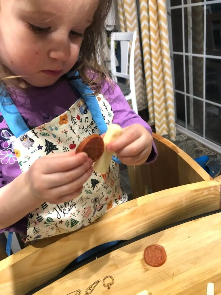 Toddler assembling snails from mozzarella sticks and pepperoni for snail pizza cooking activity.