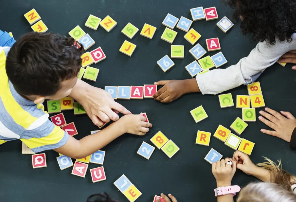 children spelling out PLAY using wooden letter blocks, demonstrating cooperative play, a stage of play.