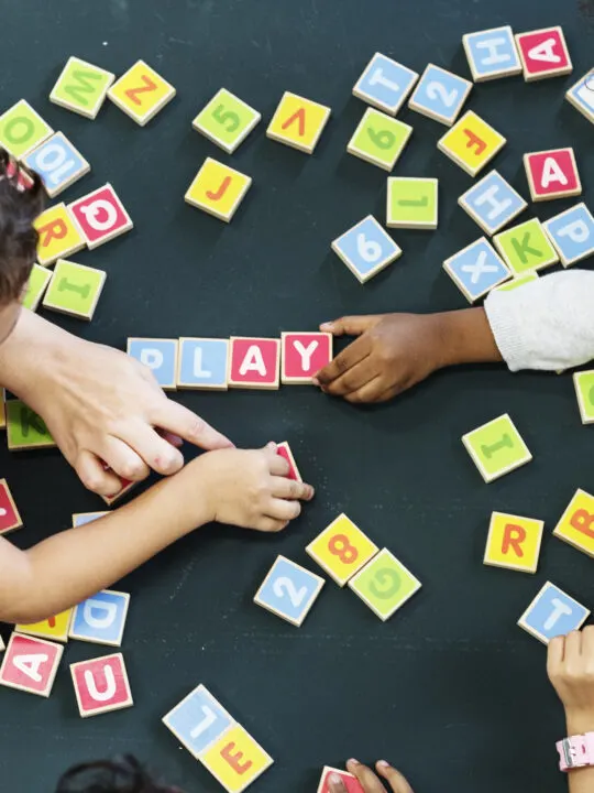 children spelling out PLAY using wooden letter blocks, demonstrating cooperative play, a stage of play.
