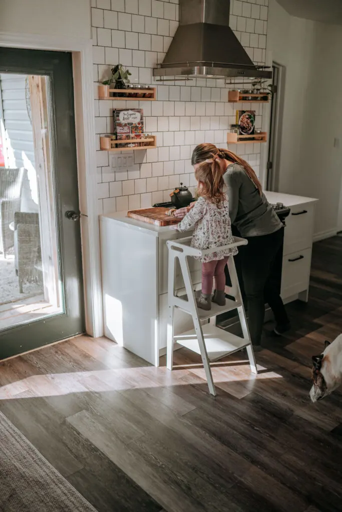 Image of child standing on Montessori learning tower in the kitchen with her mother.